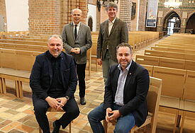 Georg Schäfer von der VR-Bank Schleswig-Mittelholstein und Piet Lassen probieren die gespendeten Stühle aus. Im Hintergrund Bischof Gothart Magaard und Pastor Michael Dübbers.