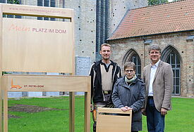 Lutz Wilhelmsen, Pröpstin Johanna Lenz-Aude und Pastor Michael Dübbers wollen mit dem Riesenstuhl auf die Stuhlpatenschaften für den Schleswiger Dom aufmerksam machen.