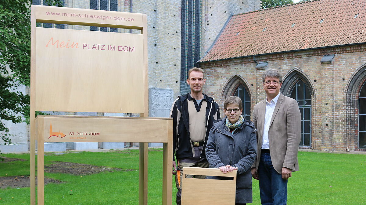 Lutz Wilhelmsen, Pröpstin Johanna Lenz-Aude und Pastor Michael Dübbers wollen mit dem Riesenstuhl auf die Stuhlpatenschaften für den Schleswiger Dom aufmerksam machen.